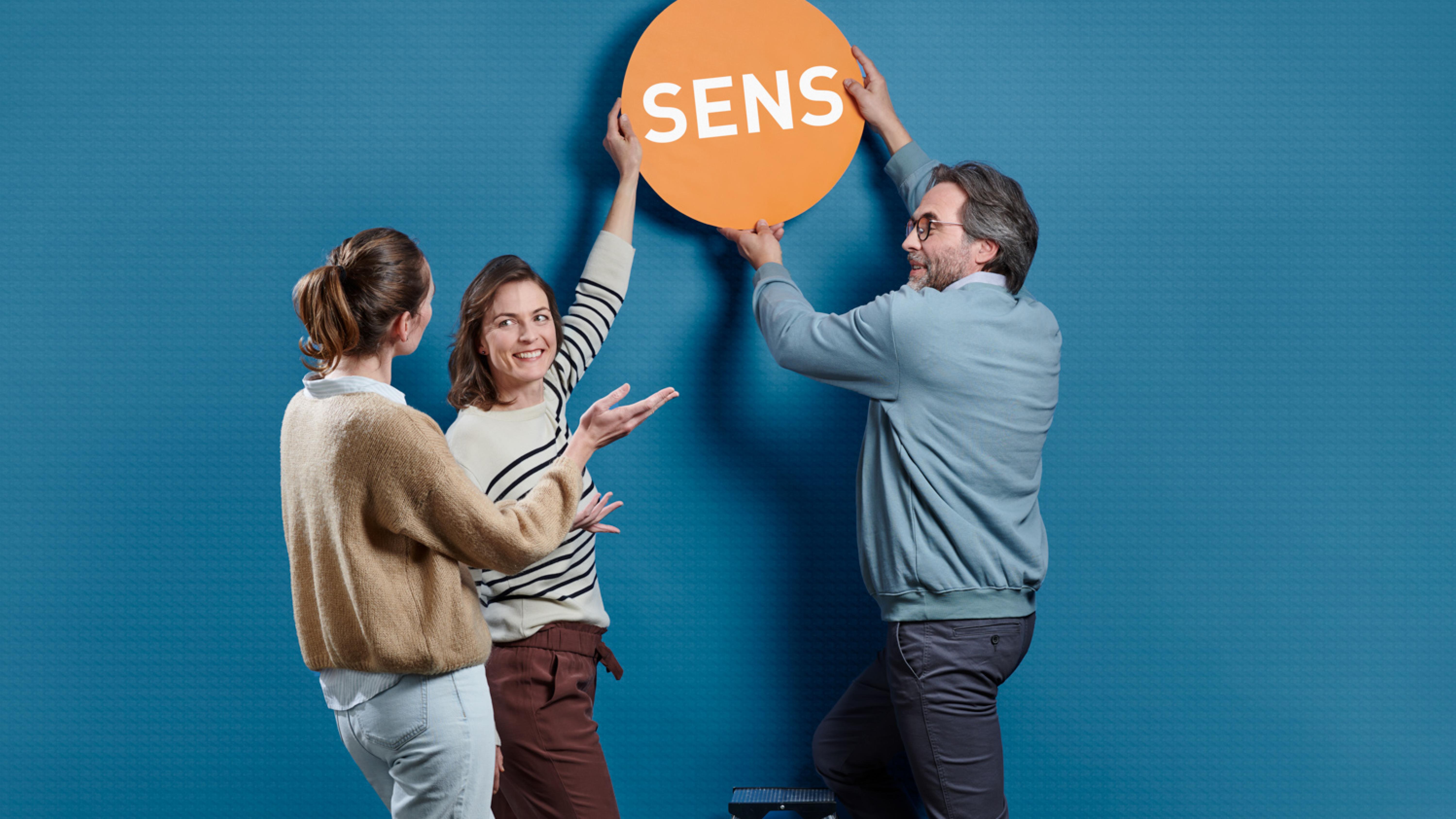 Trois personnes tiennent en l’air un panneau avec l’inscription «Sens»